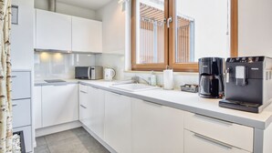 A modern kitchen with white cabinets, a built-in sink, a coffee maker, and a window with blinds. - RentPlanet - Apartament Wschodnia (Kołobrzeg)