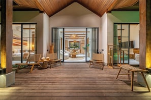 Una terraza de madera con mobiliario de mimbre, una mesa redonda en el centro y vistas a un espacio interior bien iluminado.