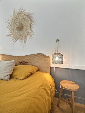 A bed with yellow bedding, a wooden headboard, a small stool, a wall-mounted lamp, and a decorative wall piece. - "Le clos du bon temps" (Lugny)