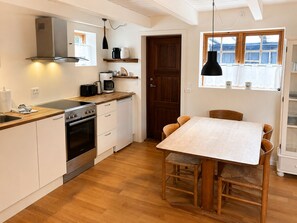 A compact kitchen with a stove, oven, and coffee maker. There is a dining table with chairs and a window with curtains. - Charming Cottage Tórshavn City Center (Tórshavn)