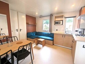 A compact kitchen with a built-in stove, a small dining table with chairs, a blue sofa, and a window providing natural light. - Cozy mobile home - 6 guests (Saint-Hilaire-de-Riez)