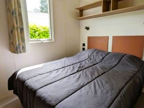 A single bed with a grey bedspread in a room with a window, a wooden shelf, and a white wardrobe. - Mobile home with TV and terrace - 4 guests (Saint-Hilaire-de-Riez)