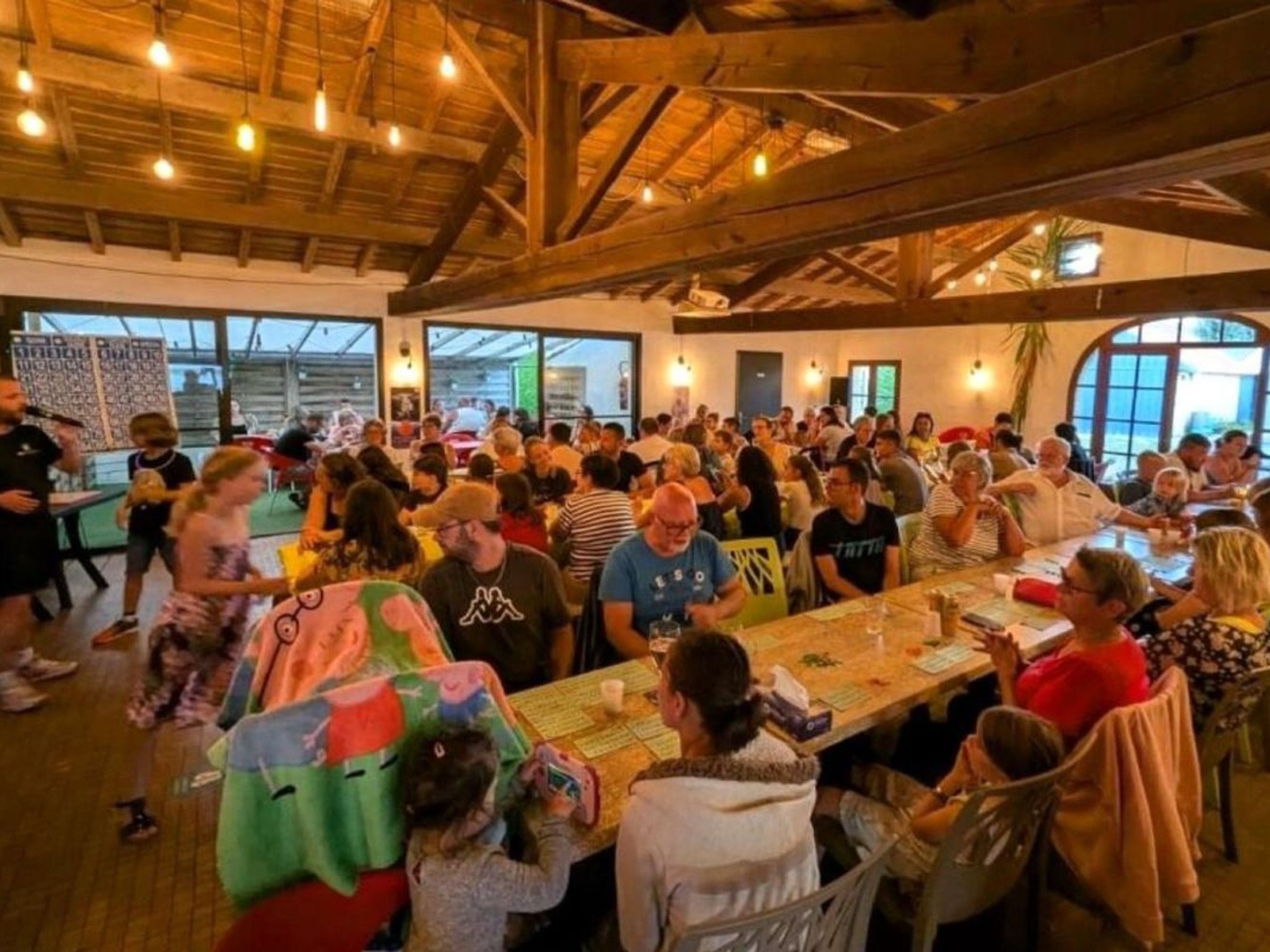 Een drukke binnenruimte met houten balken in het plafond, tafels voor groepen en grote ramen die natuurlijk licht binnenlaten.