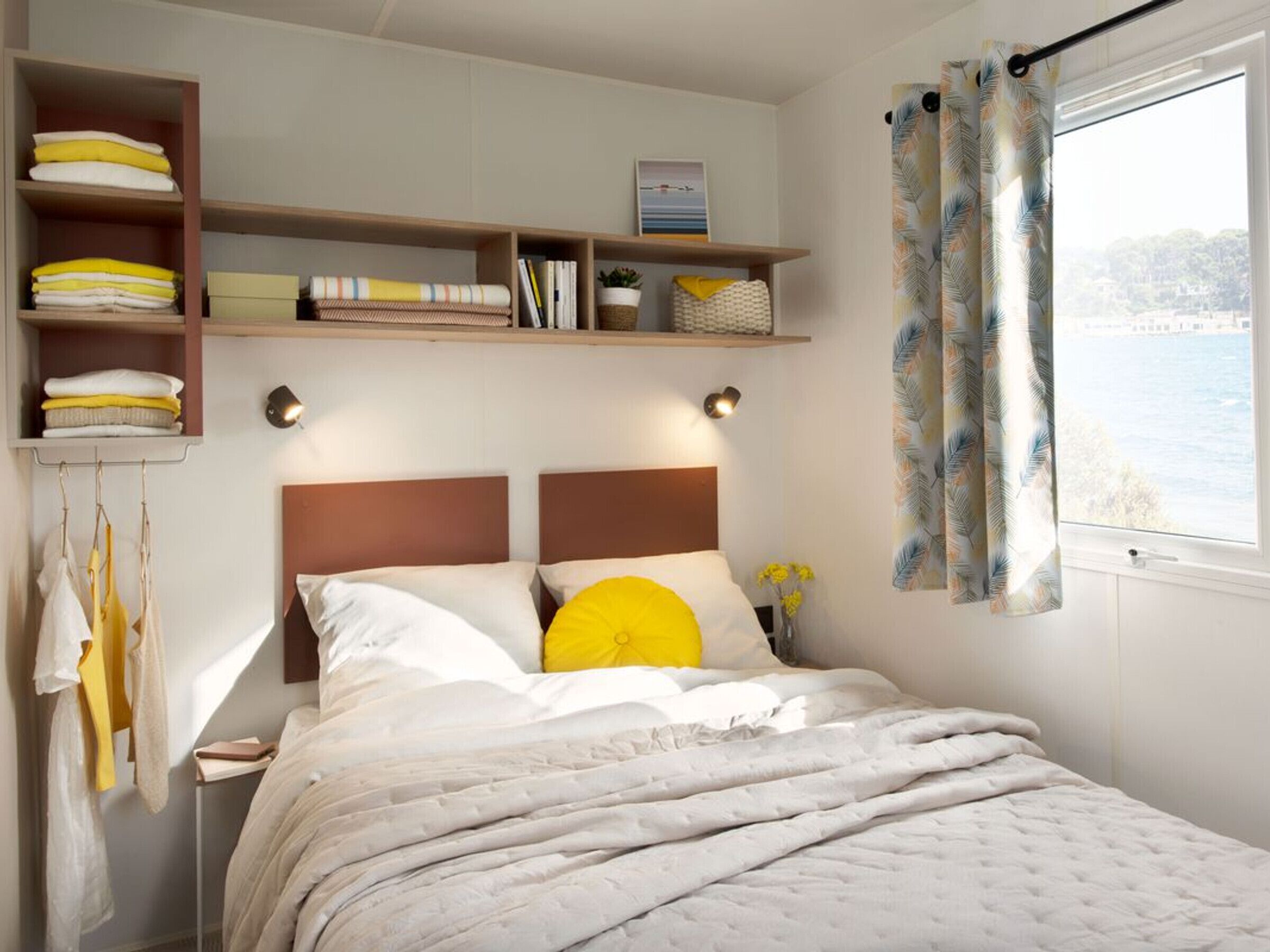 A bedroom with a bed, a shelf with folded towels, a bedside table with a lamp, and a window with curtains.