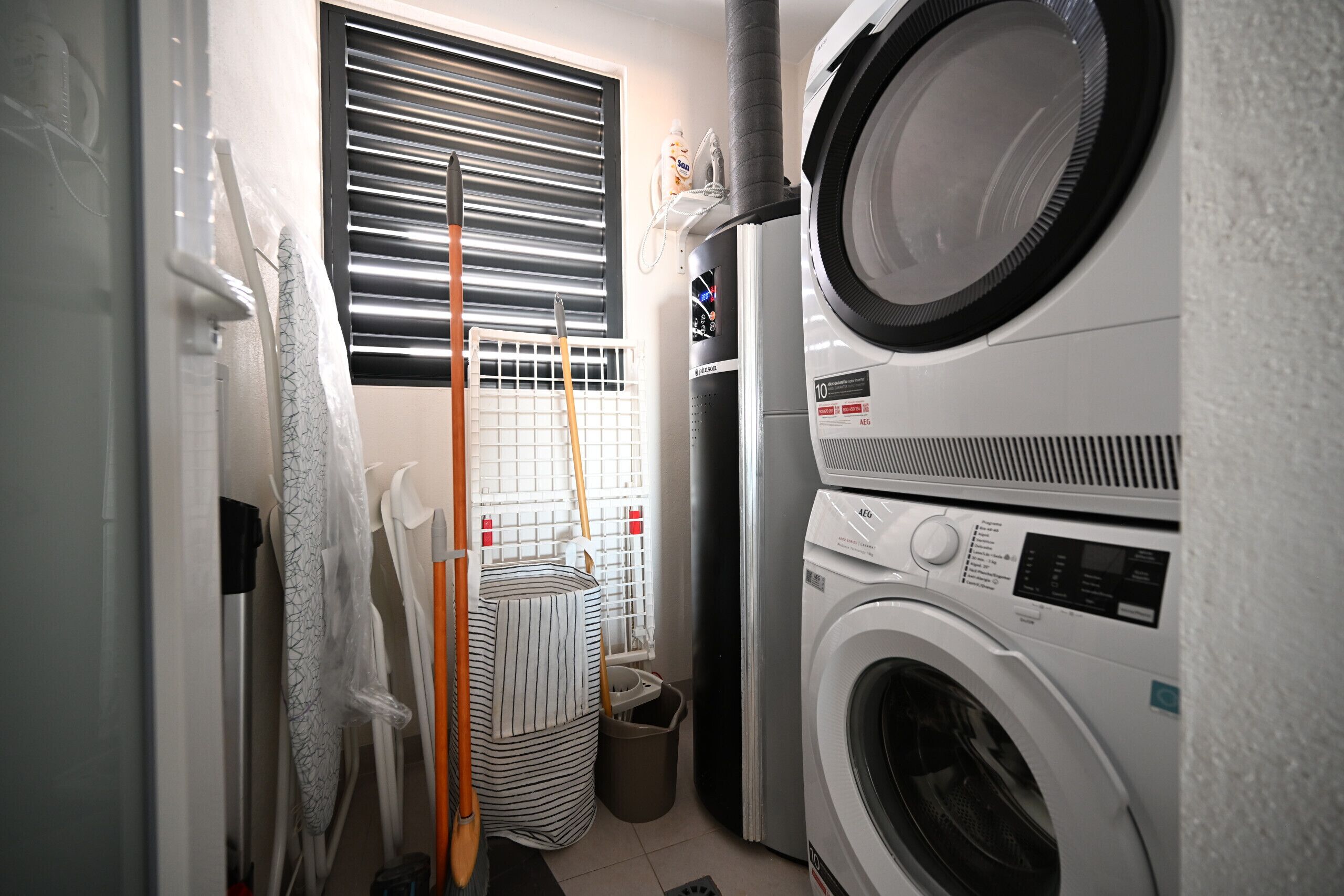 A laundry room with stacked washers and dryers, a drying rack, and a window with blinds.