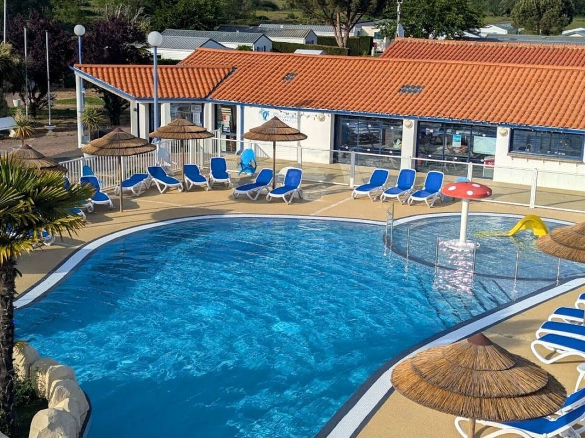 Een zwembad met blauw water, omringd door ligbedden en parasols, en een gebouw met een rood dak op de achtergrond.
