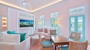 A room with a white sofa, teal cushions, a wooden table, wicker chairs, and a TV mounted on the wall. - Luxury Seaside Home Steps to Private Beach Access, Renovated, Walk to Town (Santa Rosa Beach)