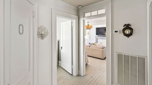 Ein Flur mit weißen Wänden, einer Tür mit einer Nummer, einem Wanddekor, einer Chandelieber, einem Thermostat und einem Lüftungsgitter.
