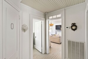 Un couloir aux murs blancs, une porte portant un numéro, un élément décoratif mural, un lustre, un thermostat et une grille d’aération.