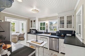 Une cuisine moderne avec des meubles blancs, un plan de travail noir, un coin repas avec des chaises en osier et une vue sur la verdure à l’extérieur.