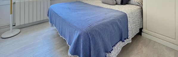 A bedroom with a bed covered by a blue blanket, a white radiator, and a round white table. - Charming apartment with AC, parking - 4 guests (Platja d'Aro)