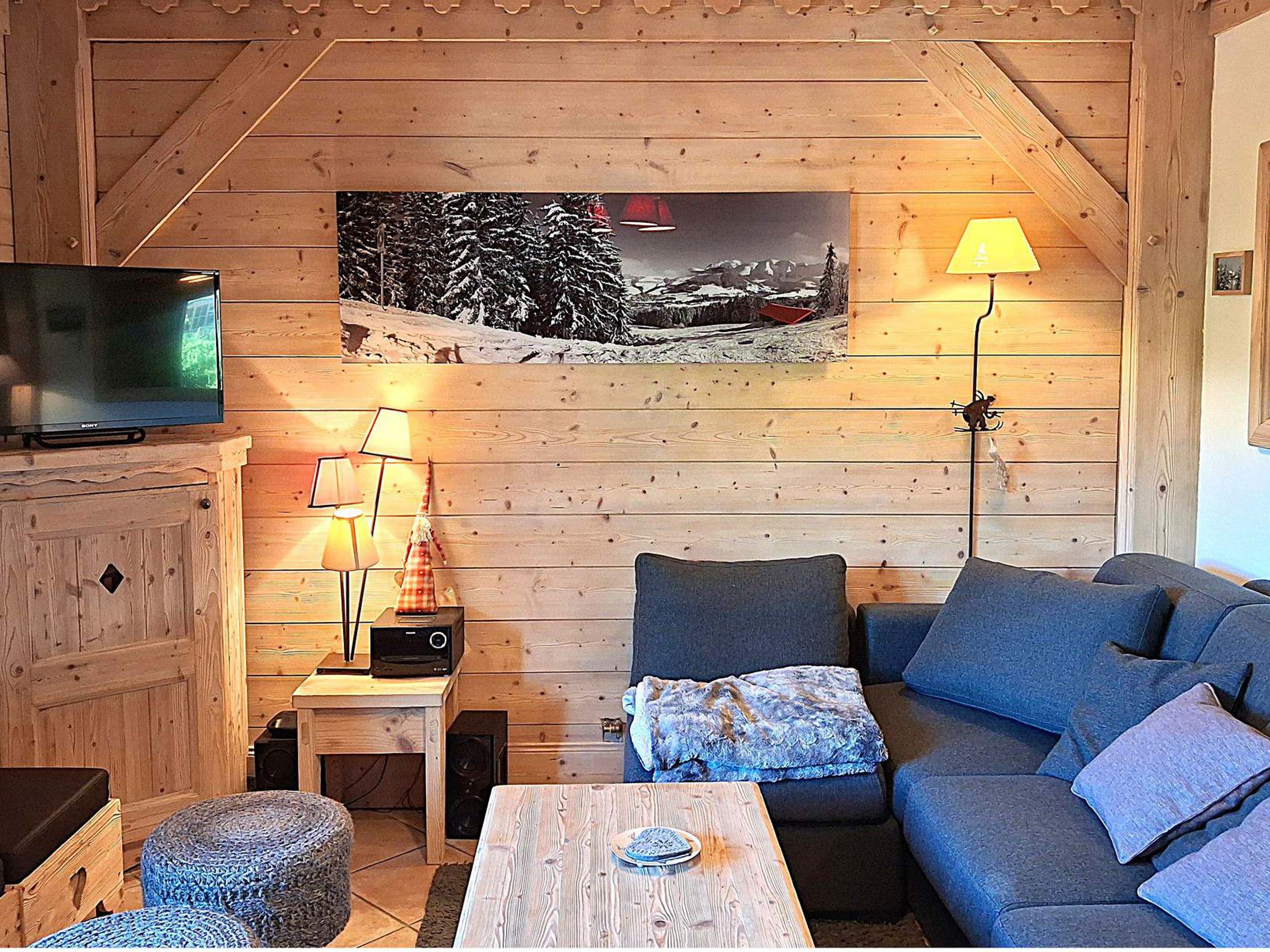 A cozy cabin interior with a wooden ceiling, a sofa, a table, a TV, and a wall-mounted lamp.