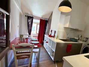 Une cuisine compacte équipée d’un lave-linge, un coin repas avec une table rouge et une fenêtre avec des rideaux.