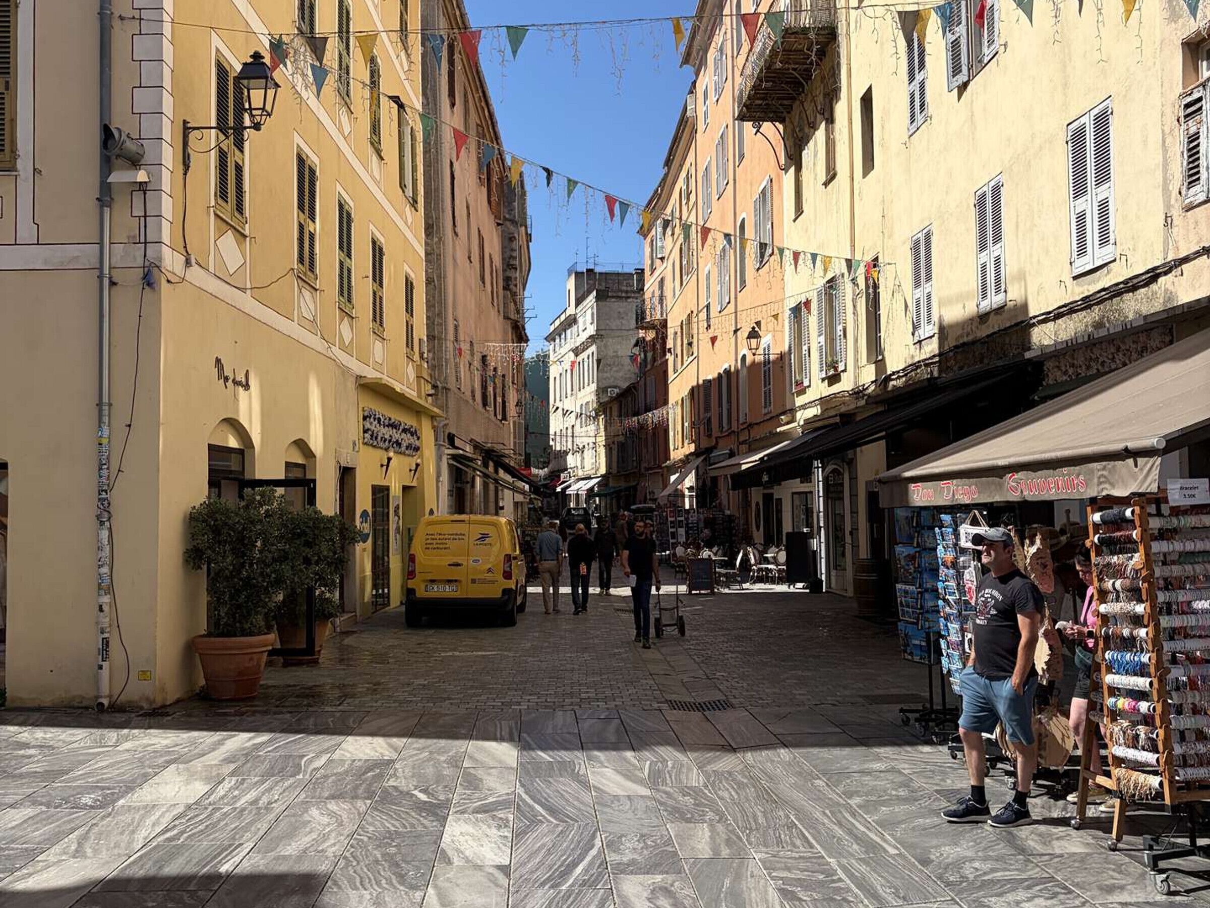 Une rue étroite, bordée de bâtiments des deux côtés, avec un fourgon jaune garé à gauche et un magasin dont l’enseigne indique « Spigoli ».