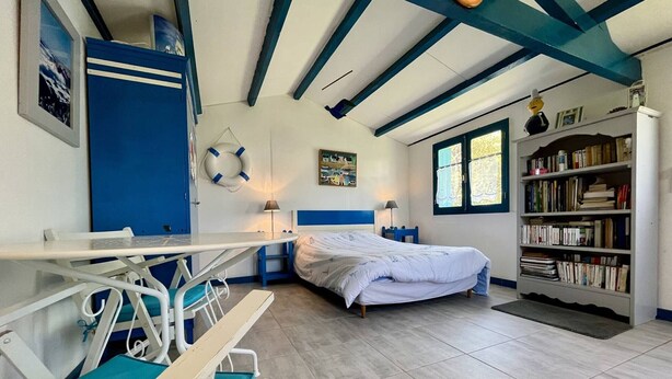 Ein Zimmer mit einem Bett, einem Schreibtisch mit Stuhl, einem Bücherregal, einem Fenster und einer Decke mit freiliegenden Balken.