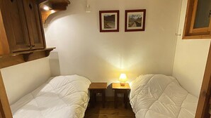Een kleine kamer met twee eenpersoonsbedden, een tafeltje met een lamp en twee ingelijste foto’s aan de muur.