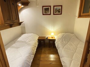 Ein schmaler Raum mit zwei Einzelbetten, einem kleinen Tisch mit Lampe und zwei gerahmten Bildern an der Wand.