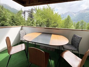 Ein Balkon mit rundem Tisch und vier Stühlen, der auf eine gebirgige Landschaft mit Bäumen und einem Gebäude blickt.
