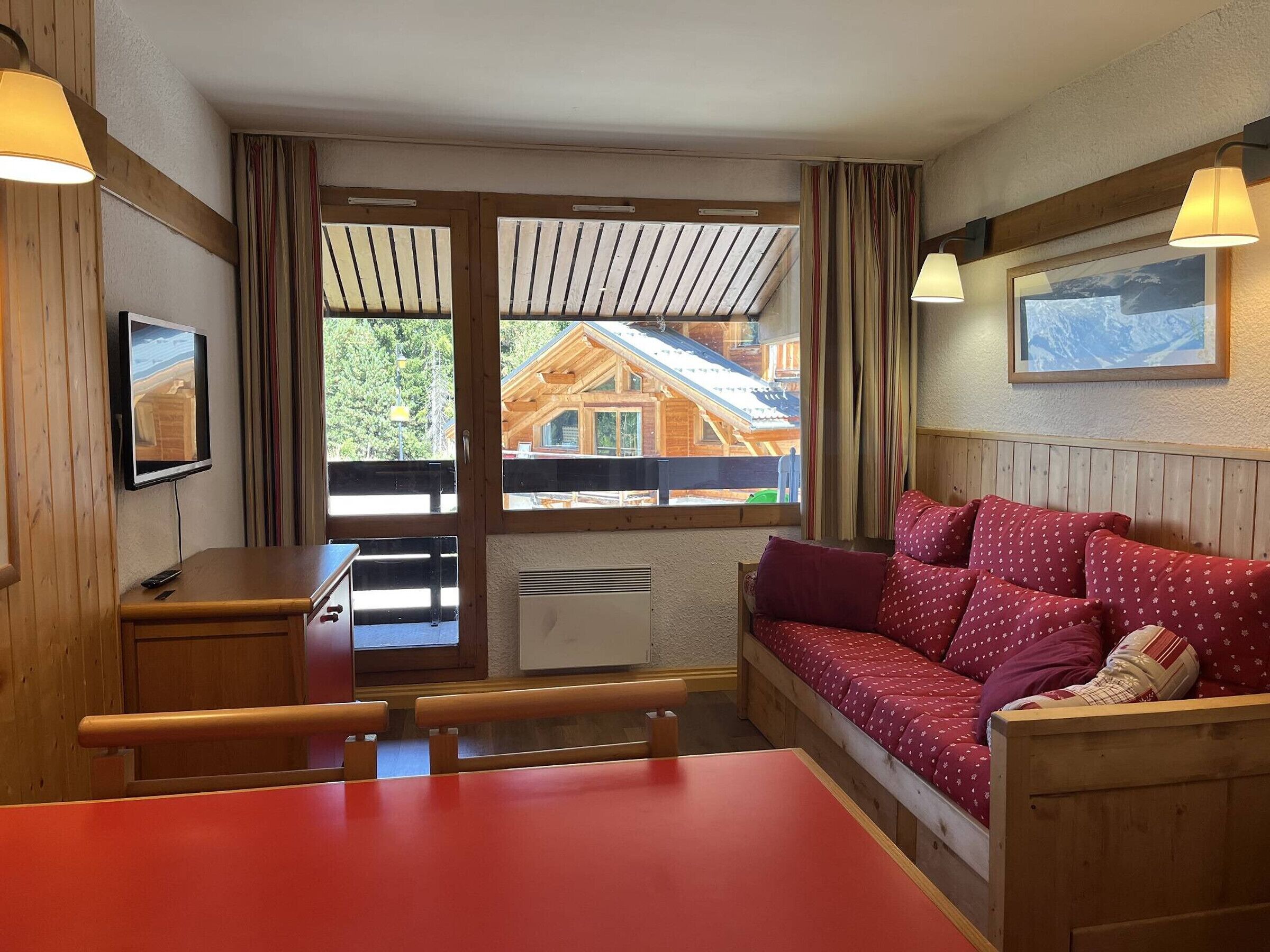 A cozy room with a wooden bench, a red sofa, a TV, and a window with a view of a wooden cabin.