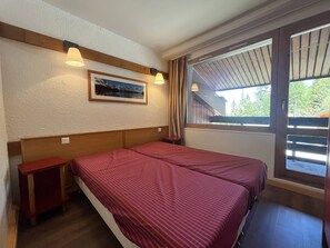 Ein Zimmer mit zwei Betten, einem Holzkopfende, einem kleinen Tisch, einem Fenster mit Vorhängen und einem gerahmten Bild an der Wand.