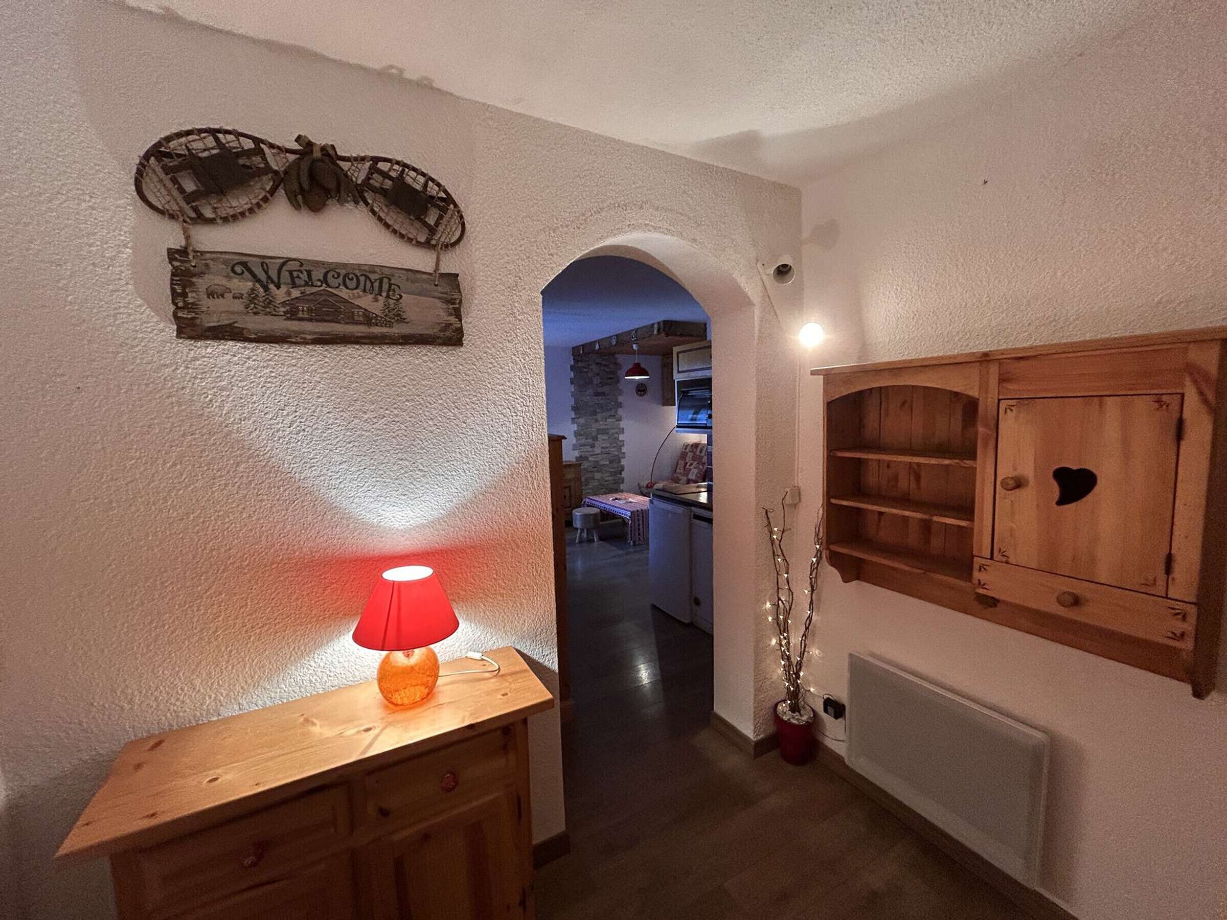 A hallway with a wooden cabinet, a red lamp, a welcome sign, and a view into a kitchen area.