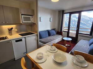 A compact hotel room with a kitchenette, a sofa bed, a dining table with chairs, and a view of a snowy landscape. - Apartment with view - 8 guests (Avoriaz)
