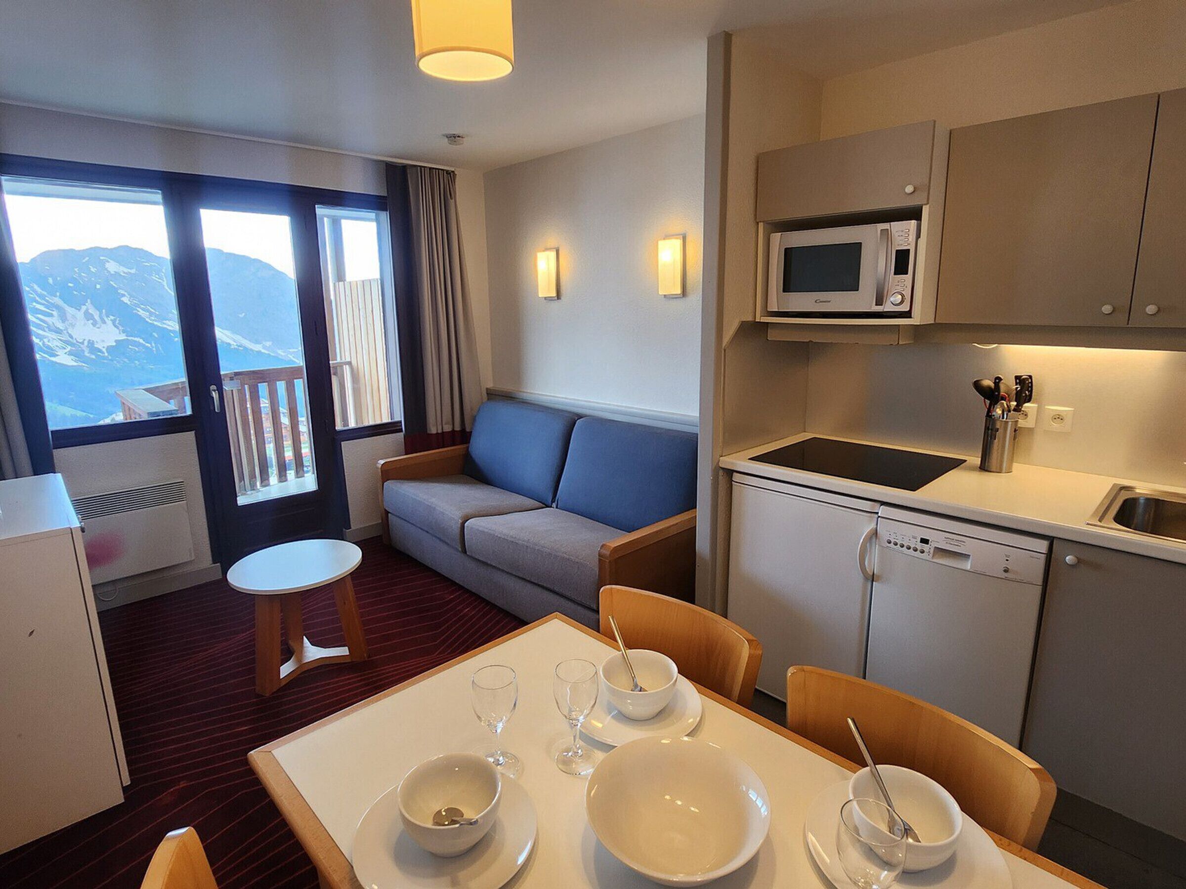 A compact hotel room with a balcony, a sofa, a dining table with bowls and glasses, a microwave, and a dishwasher.
