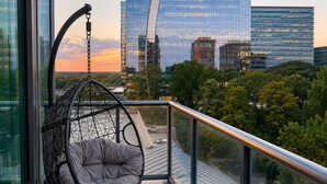 Een balkon met een hangstoel, uitzicht op een stadsgezicht met moderne gebouwen en een heldere lucht.