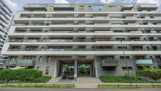 Un edificio residencial de varios pisos con balcones, un acceso cubierto y un jardín delantero ajardinado.
