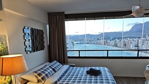 Habitación de hotel con cama, escritorio, silla, lámpara y vistas a una ciudad y montañas a través de amplios ventanales.