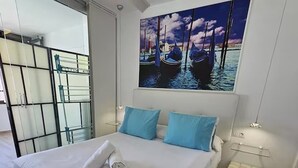 Habitación de hotel con cama, un cuadro de barcos en el agua, una puerta de vidrio y una mesita de noche pequeña con una lámpara.
