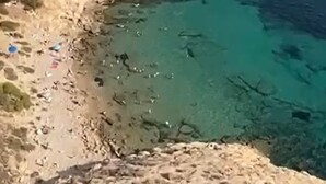 Un paisaje costero con aguas azules cristalinas, terreno rocoso y una bandera en un acantilado.