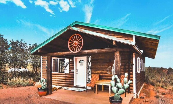 Eine rustikale Blockhütte mit Veranda, einem Rollzeichen, einer Kaktusskulptur und einem Schild mit der Aufschrift 'RANCH'.