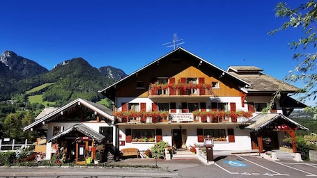 A hotel with a restaurant and bar, a parking area, and mountainous scenery in the background.. Le plein soleil