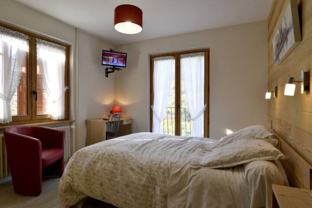 Un dormitorio con cama, una silla roja, un televisor montado en la pared y una ventana con cortinas.