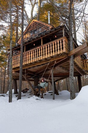 Une cabane en bois perchée dans un arbre, dotée d’un balcon et entourée d’arbres et de neige.