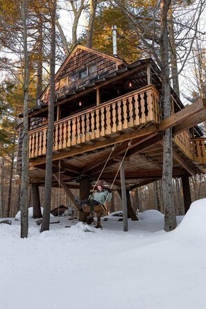 Une cabane en bois perchée dans un arbre, dotée d’un balcon et entourée d’arbres et de neige.