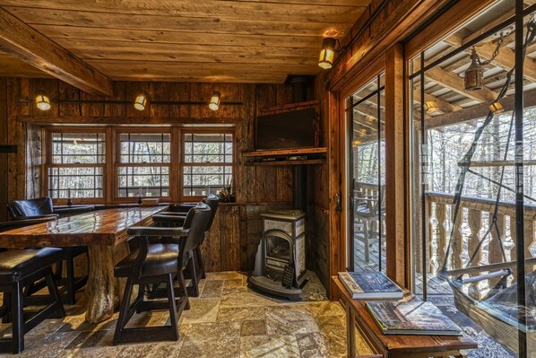 Un intérieur rustique en bois, avec une grande table à manger, des fauteuils en cuir, une cheminée et de grandes fenêtres donnant sur l’extérieur.