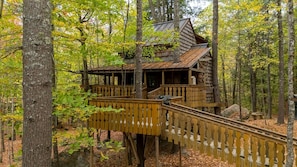 A wooden cabin with a wooden deck and stairs leading up to it, surrounded by trees.