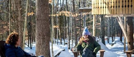 Two people sitting on wooden chairs by a fire pit in a snowy forest setting.
