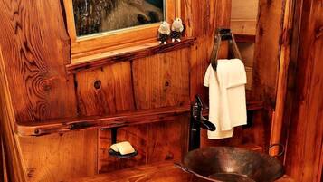A rustic wooden cabin interior with a wooden sink, a window with a view of a tree, and a shelf with two plush toys.