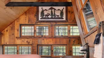 Une pièce chaleureuse dans un chalet, avec un plafond en pente, des murs en bois, un lit recouvert d’une couverture rose, un lavabo en bois et de nombreuses fenêtres.
