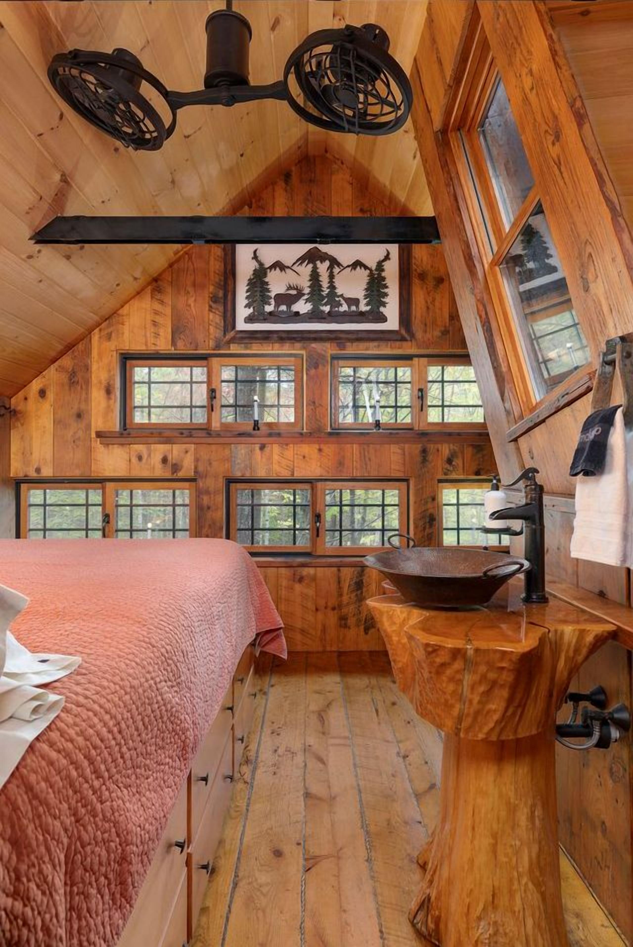 Ein gemütliches Blockhauskabinenzimmer mit schrägen Wänden, Holzwänden, einem Bett mit rosa Decke, einem Holzwaschbecken und mehreren Fenstern.