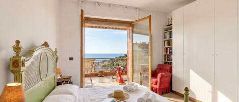 A bedroom with a bed, a nightstand, a lamp, a window, a balcony, a chair, and a bookshelf.