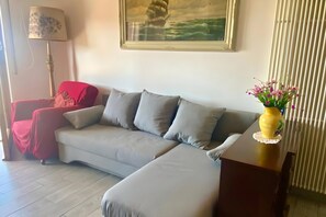 Ein Wohnzimmer mit einem grauen Sofa, einem roten Sessel, einer Lampe, einem gerahmten Bild eines Schiffes und einer Vase mit Blumen.