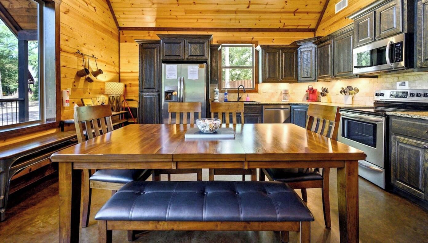 A wooden cabin interior with a dining table, chairs, a bench, a refrigerator, a microwave, and a stove.