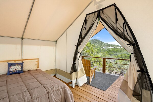 A tented room with a bed, a chair, and a view of a mountainous landscape.