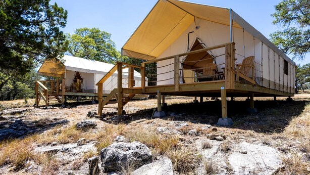 Varias cabañas de madera con techos de lona, ubicadas en un terreno rocoso y natural con árboles al fondo.