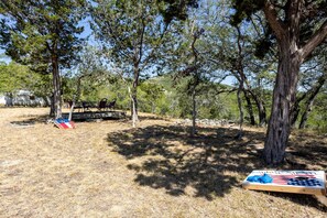 A campsite with a table and chairs under a tree, a flag on the ground, and a wooden structure in the background.
