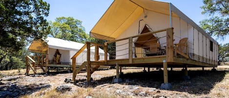 A series of wooden cabins with canvas roofs, set in a natural, rocky terrain with trees in the background.